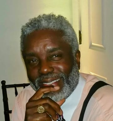 Smiling Black man with graying hair and beard, wearing a pink shirt, black suspenders, and a ring.