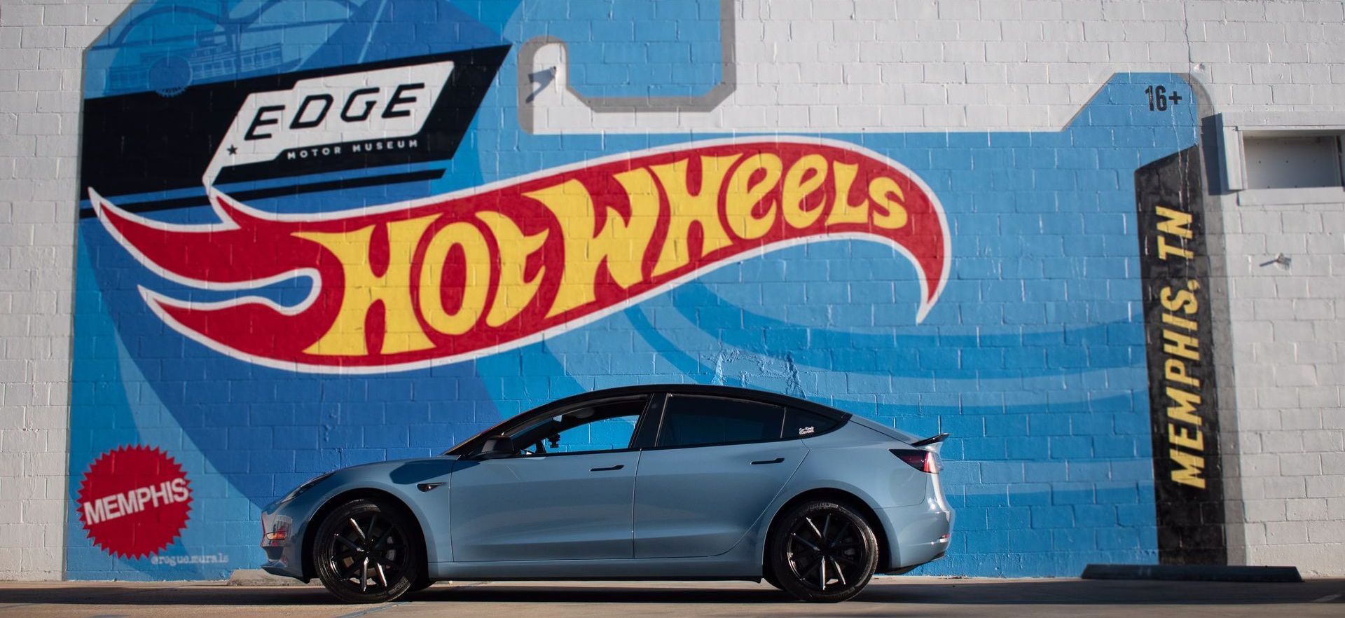 a Tesla Model 3 wrapped in hot wheels blue vinyl is parked in front of a hot wheels mural on a wall.