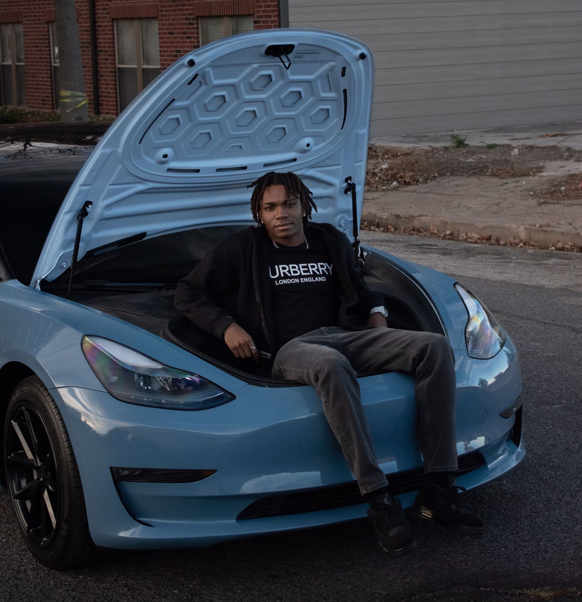 a man is sitting on the hood of a hot wheels blue tesla model 3