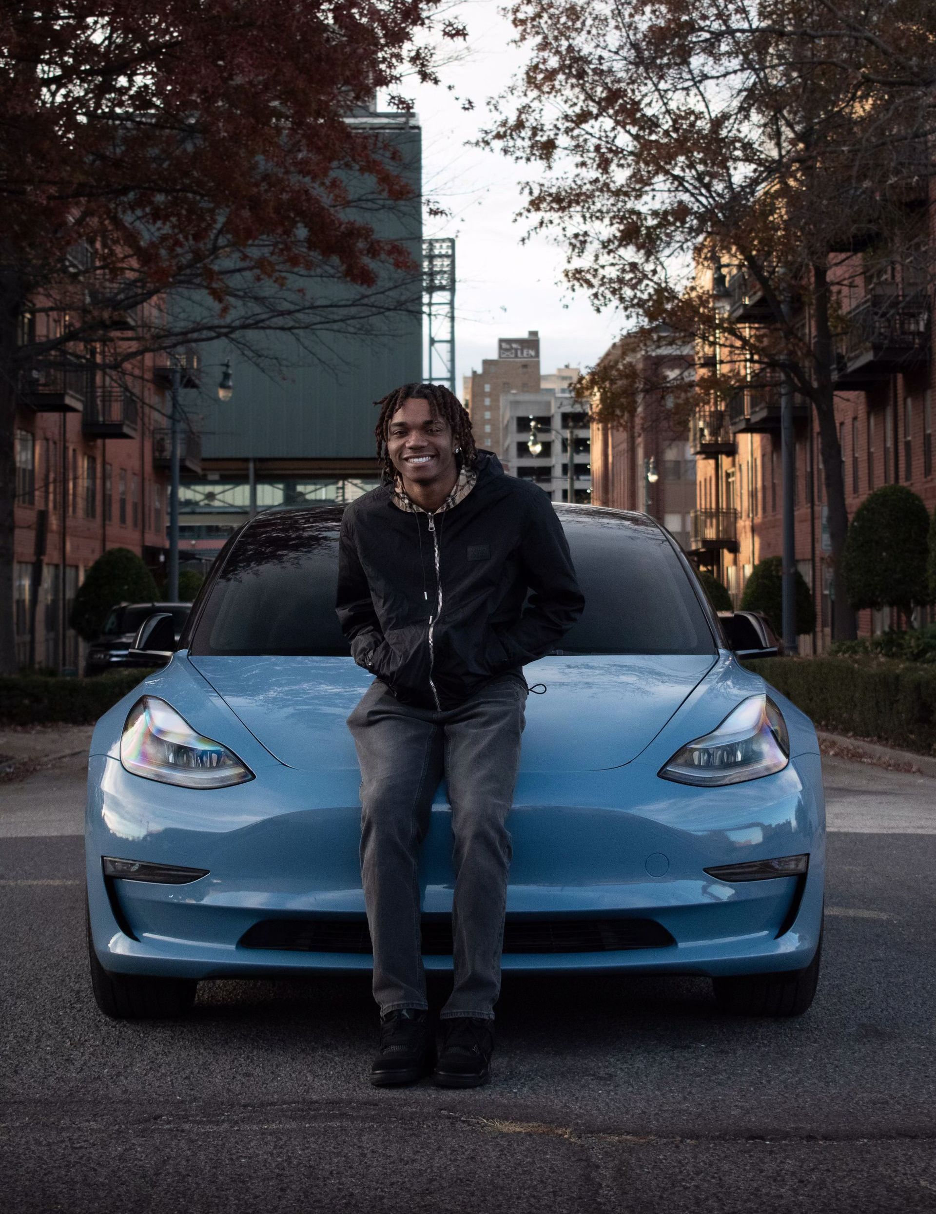 a man is sitting on the front of a blue tesla model 3 .