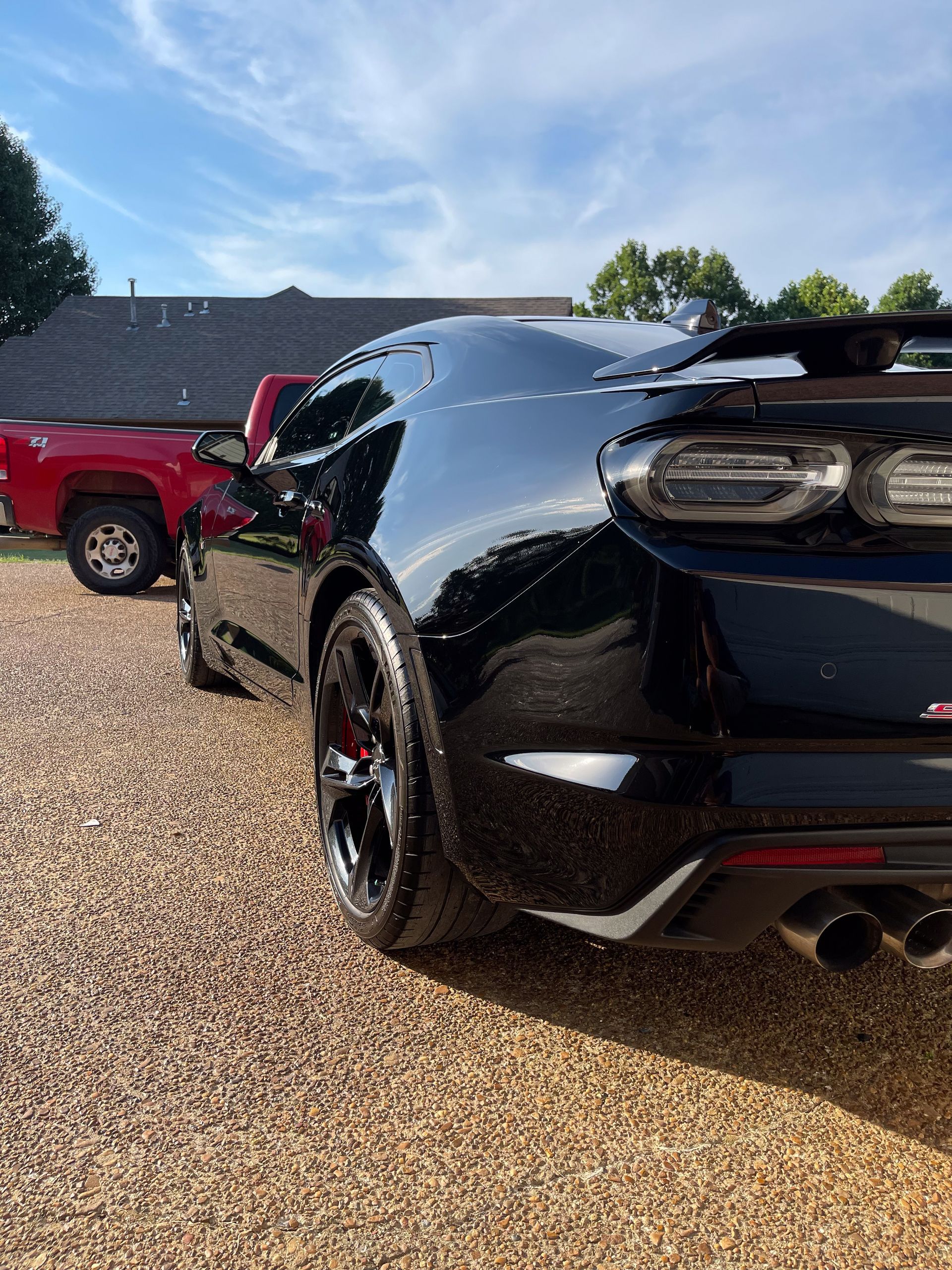a black sports car is parked in a gravel driveway next to a red truck.