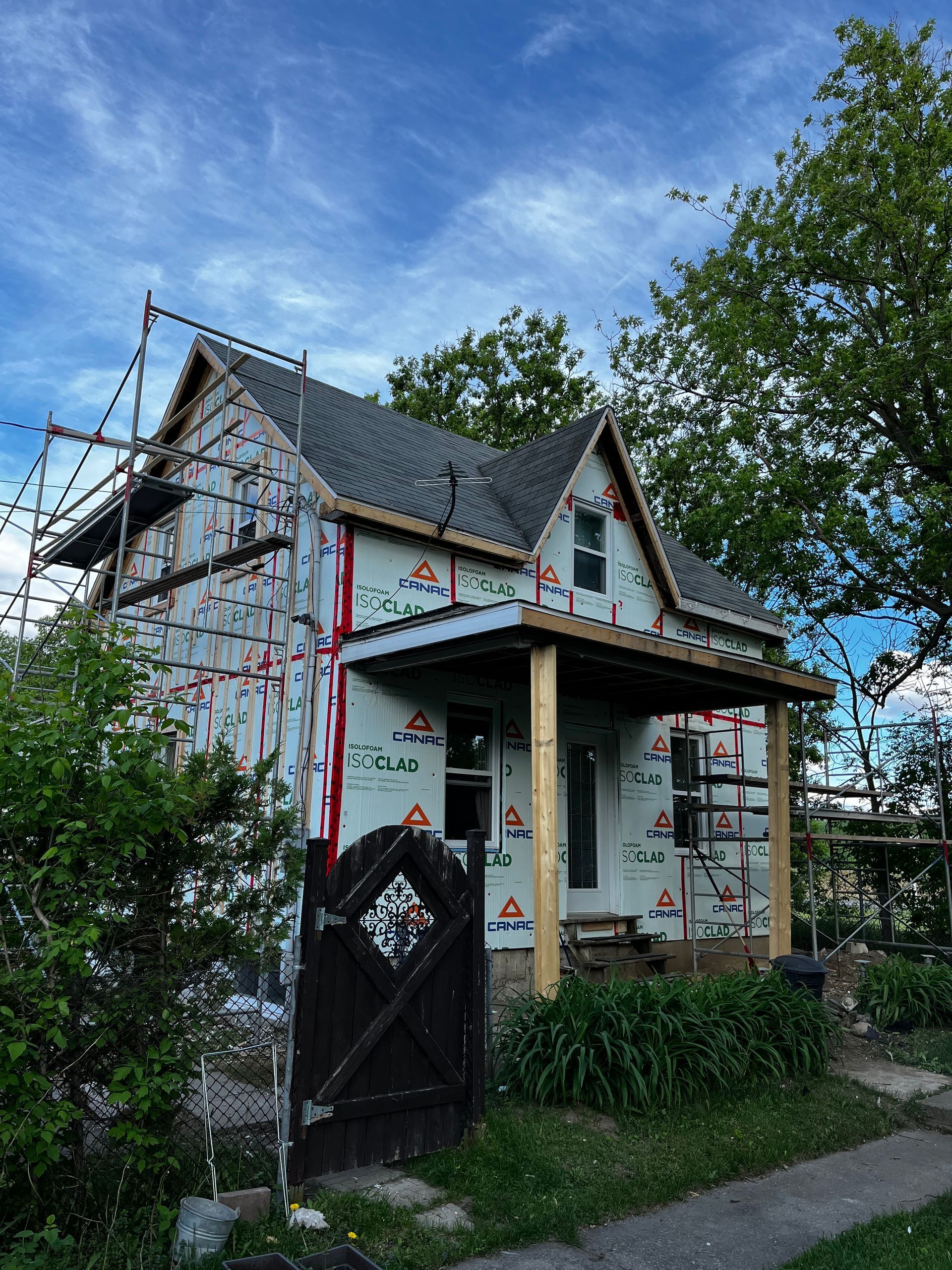 Maison en construction, recouverte d'une bâche résistante aux intempéries. Des échafaudages entourent le bâtiment, porche visible, ciel nuageux.