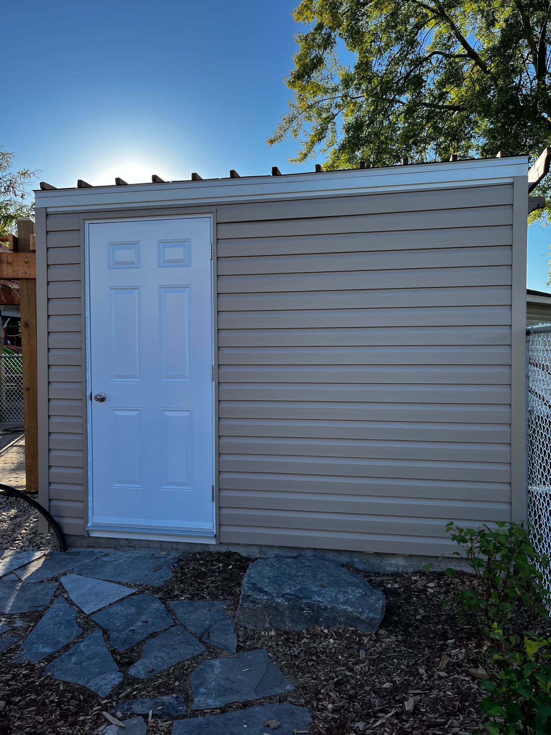 Abri beige à porte blanche, situé sur un chemin de pierre avec un ciel bleu ensoleillé en arrière-plan.