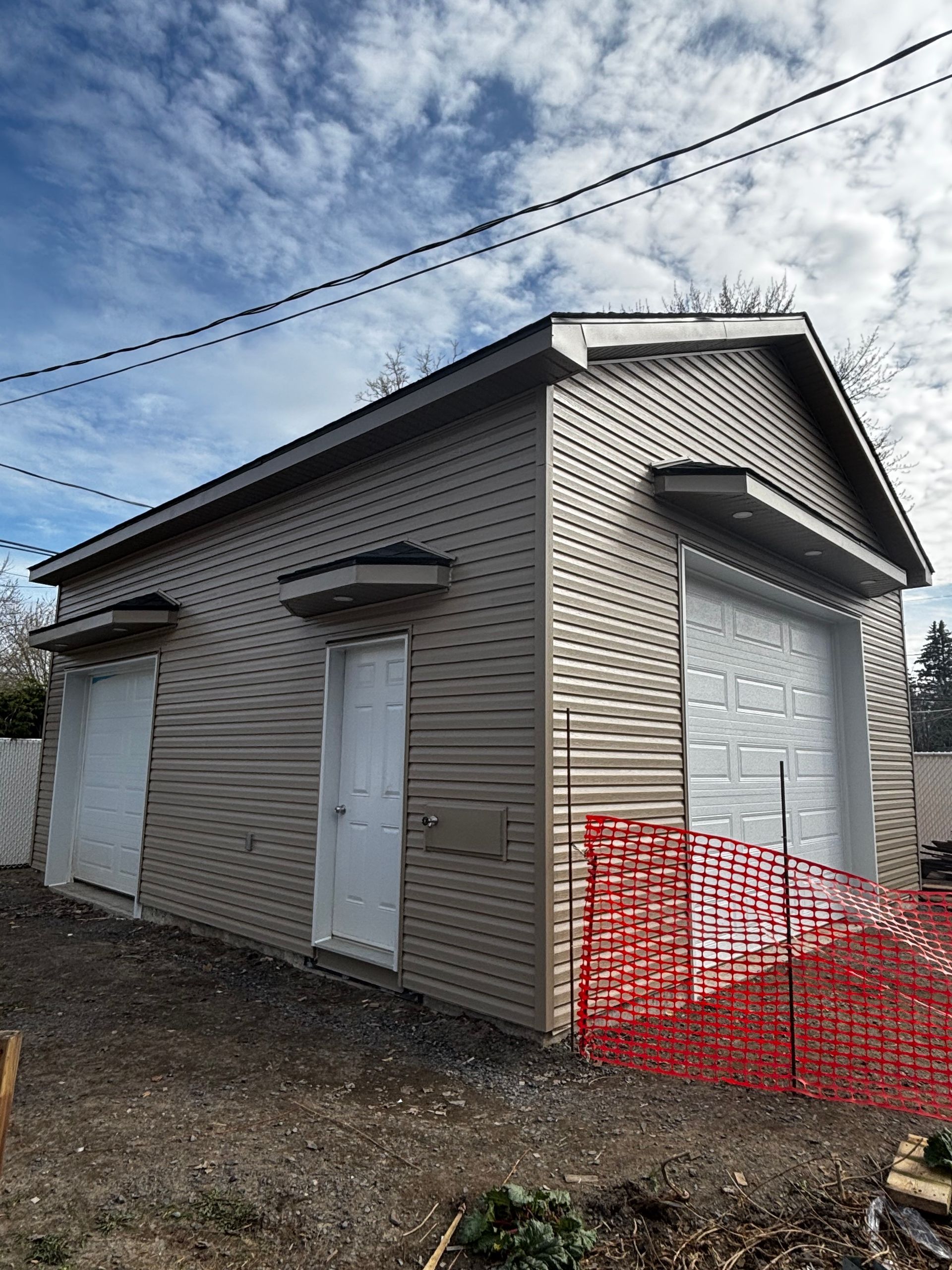 Garage beige à deux travées, portes blanches et clôture de chantier rouge sous un ciel partiellement nuageux.