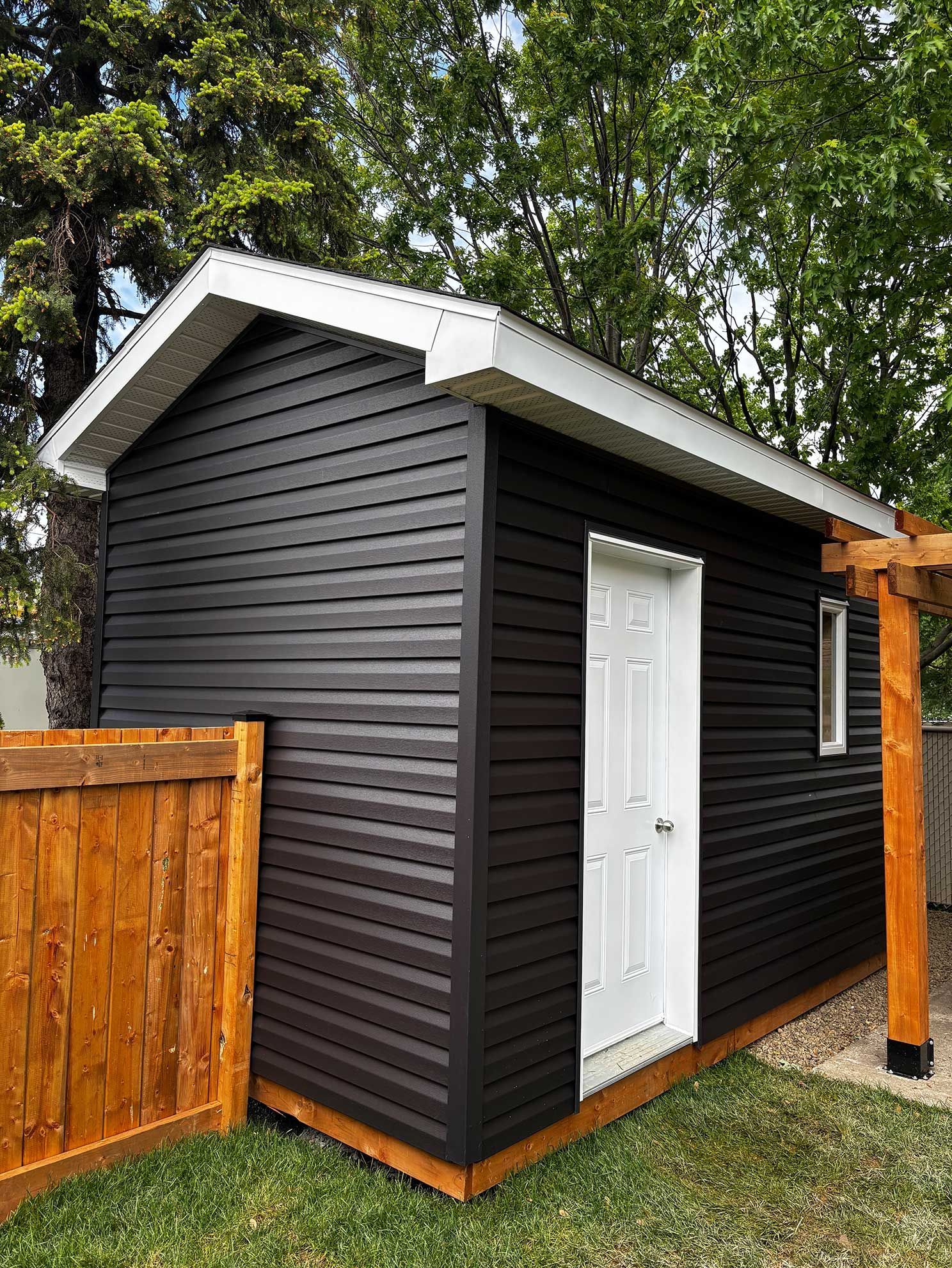 Abri de jardin noir avec boiseries et porte blanches, à côté d'une clôture en bois.