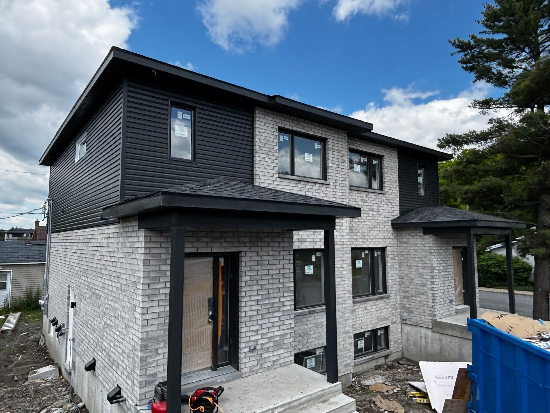 Duplex de deux étages avec façade en briques grises, bardage gris foncé, finitions noires et petites entrées couvertes sous un ciel bleu.