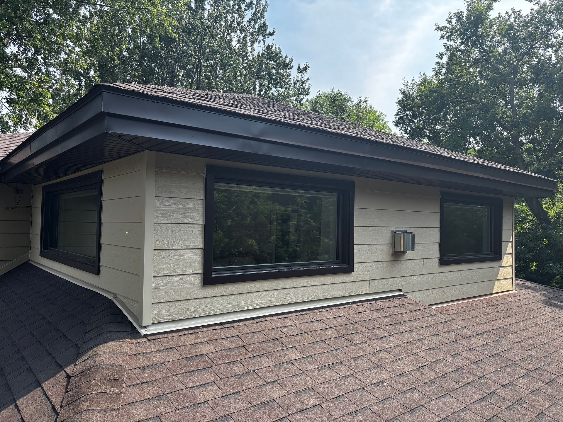 Toit marron avec lucarne, bardage beige, fenêtres à encadrement noir et petit climatiseur.