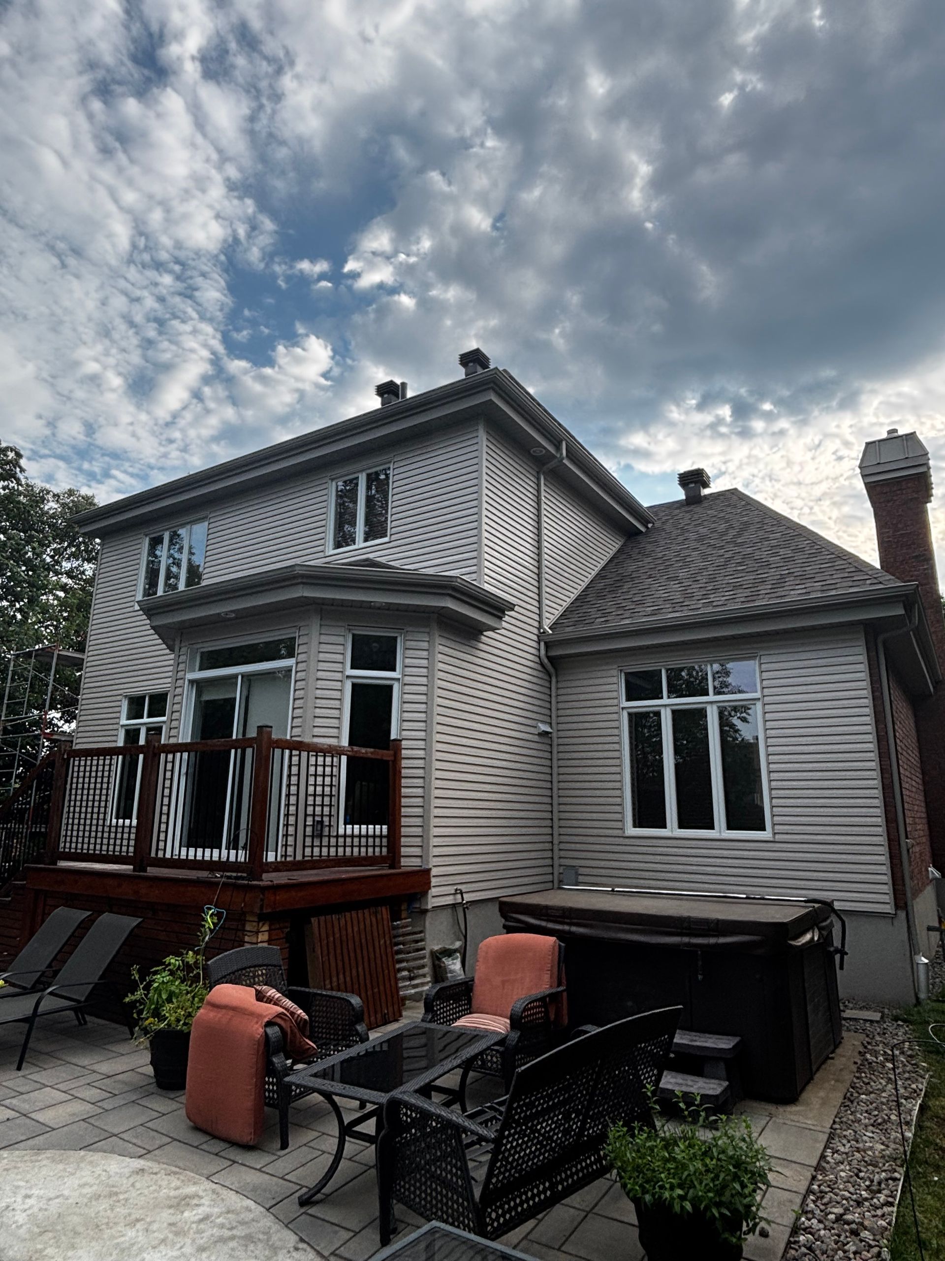 Vue arrière d'une maison à deux étages avec terrasse, mobilier de jardin et jacuzzi sous un ciel nuageux.