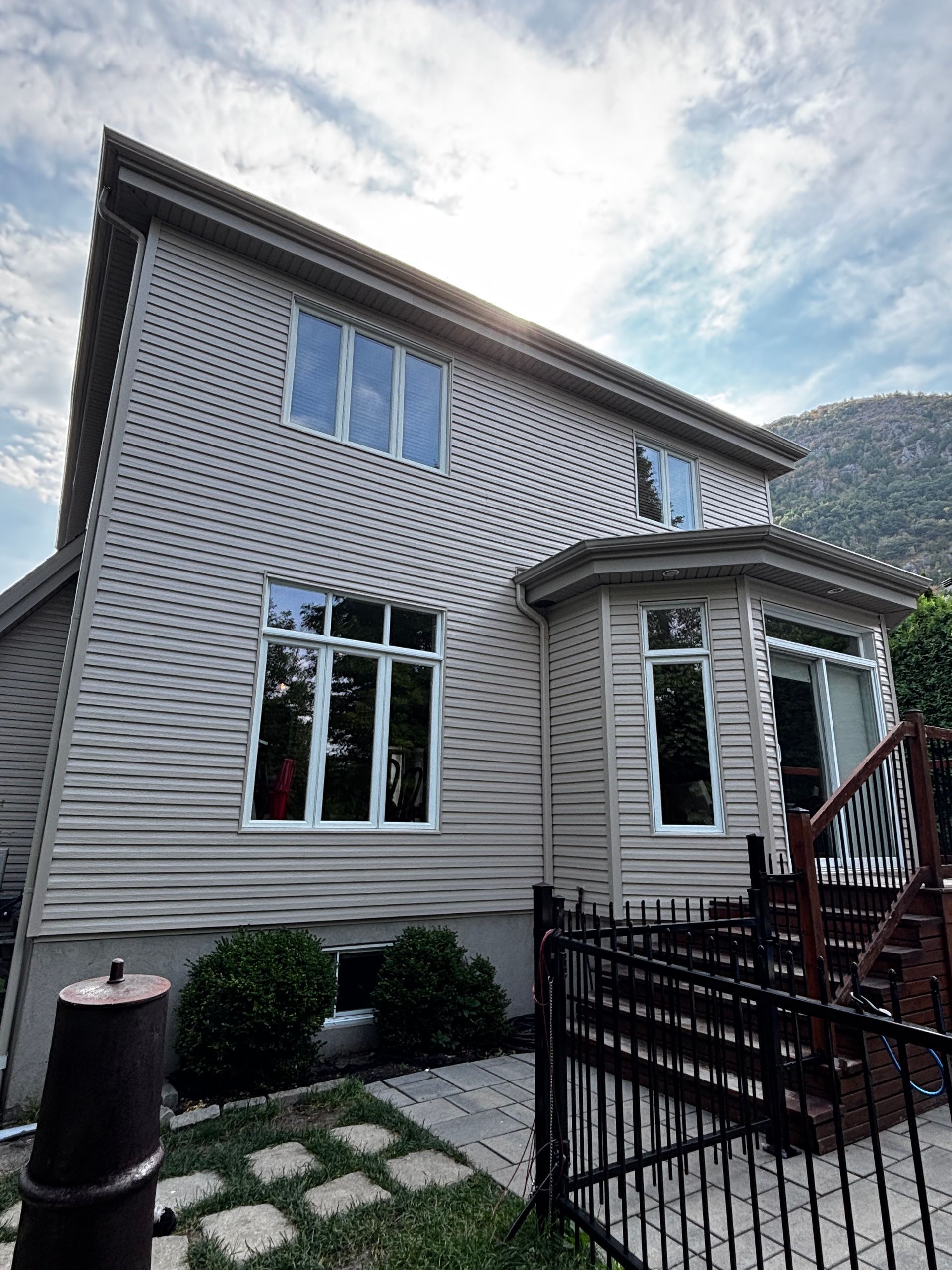 Maison beige à deux étages avec de nombreuses fenêtres et des boiseries foncées. Clôture noire, escalier et montagne visible en arrière-plan.