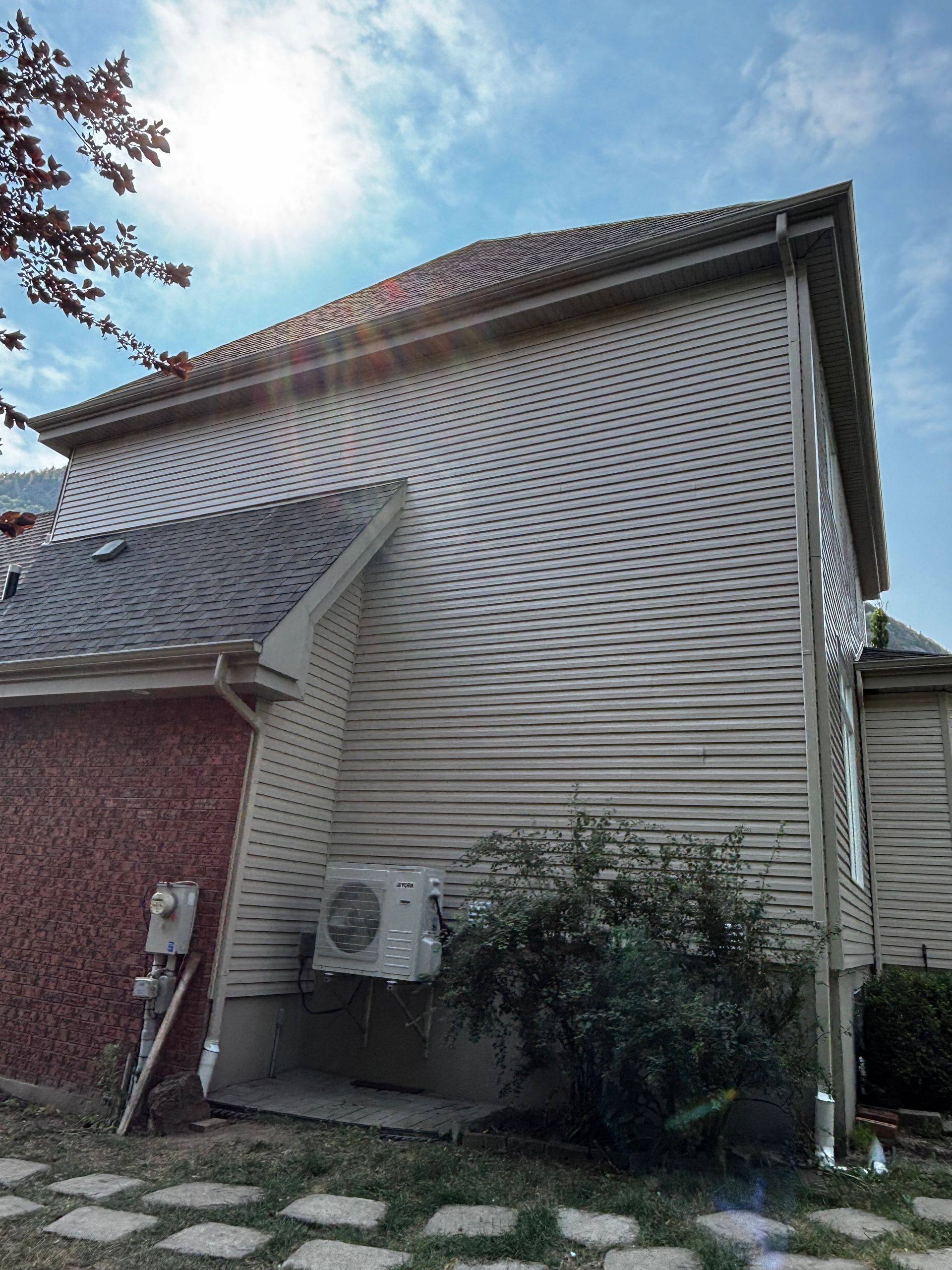 Façade d'une maison avec un revêtement extérieur clair, un climatiseur et des arbustes sous un ciel bleu.
