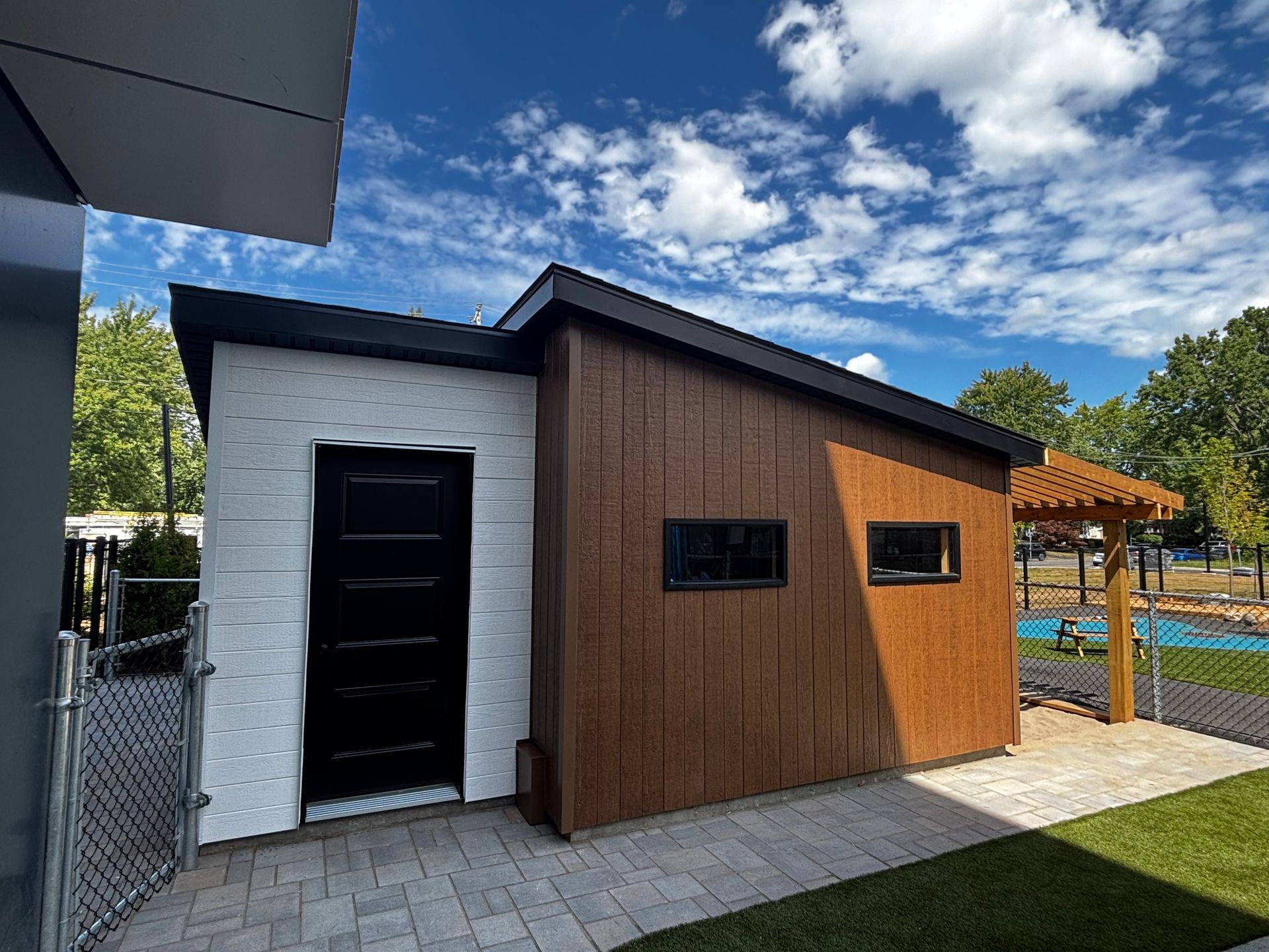Abri de jardin moderne avec bardage blanc et marron, porte noire et petites fenêtres, sur une terrasse en briques sous un ciel bleu.
