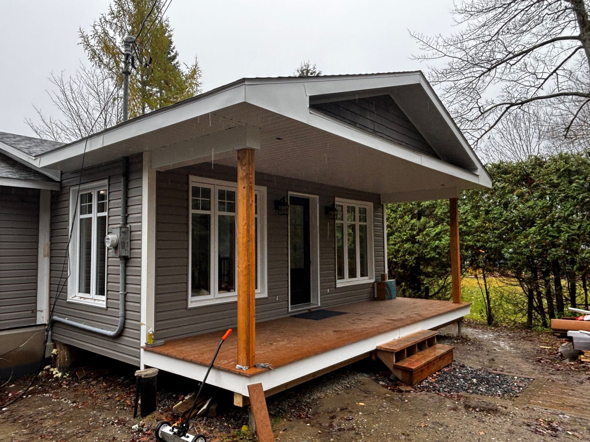Petite maison avec porche couvert ; bardage gris, boiseries blanches, porche et supports en bois, marches.