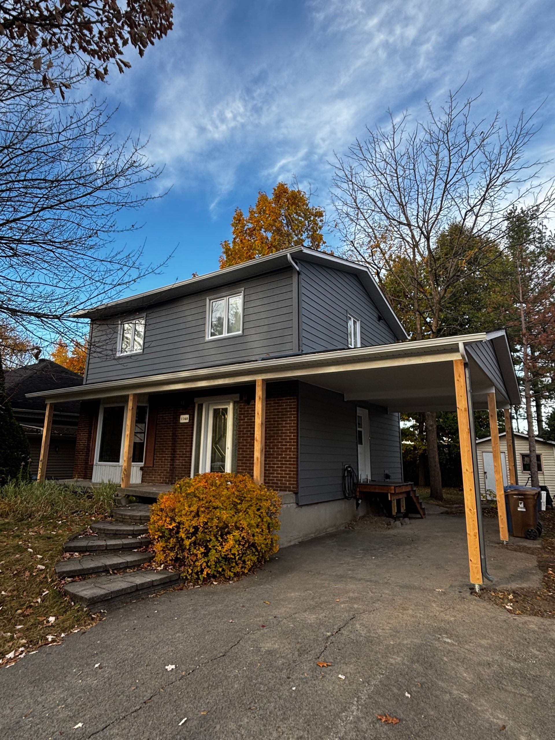 Maison grise à deux étages avec un abri voiture ; des marches en pierre mènent à la porte d'entrée, feuillage d'automne et ciel bleu.