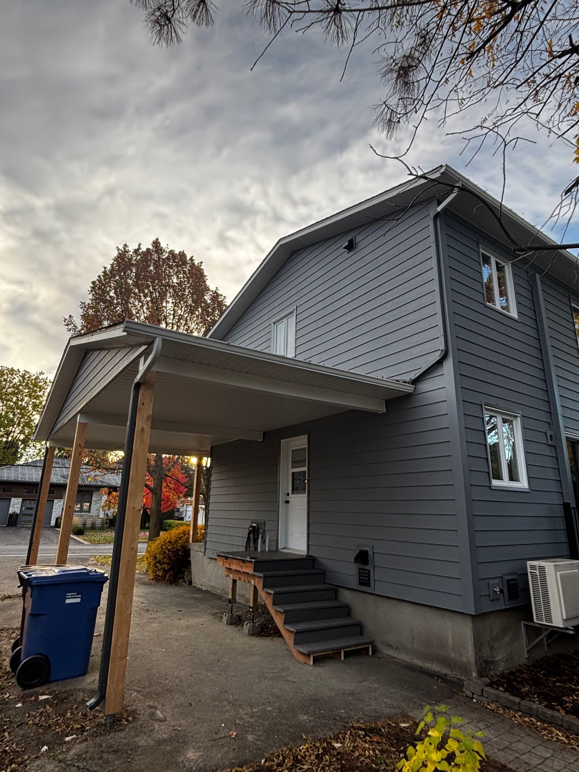 Maison grise à deux étages avec un porche et des marches menant à la porte d'entrée ; poubelle bleue visible.
