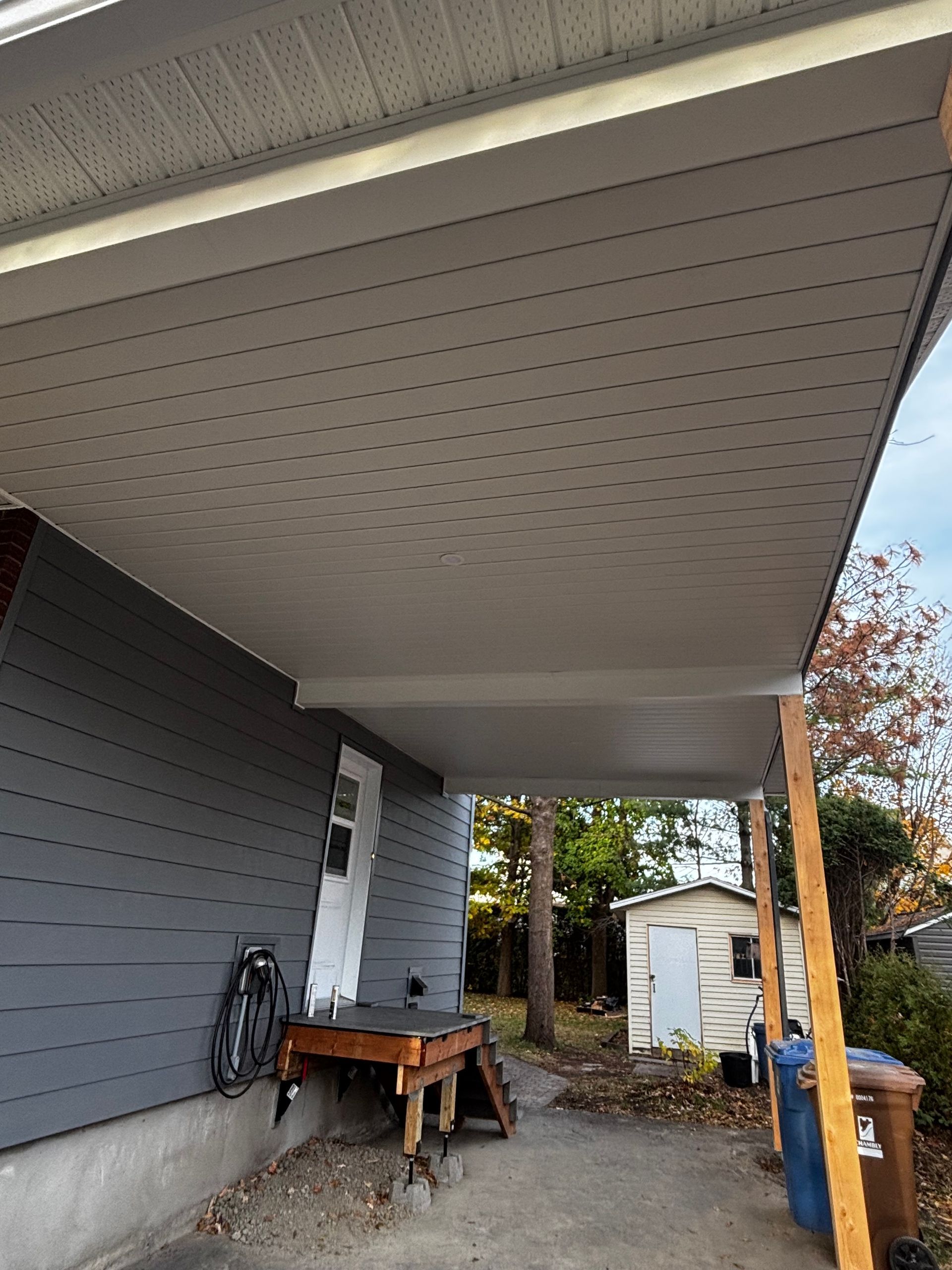 Abri de voiture gris avec plafond gris clair, attenant à une maison grise. Vue sur le jardin.