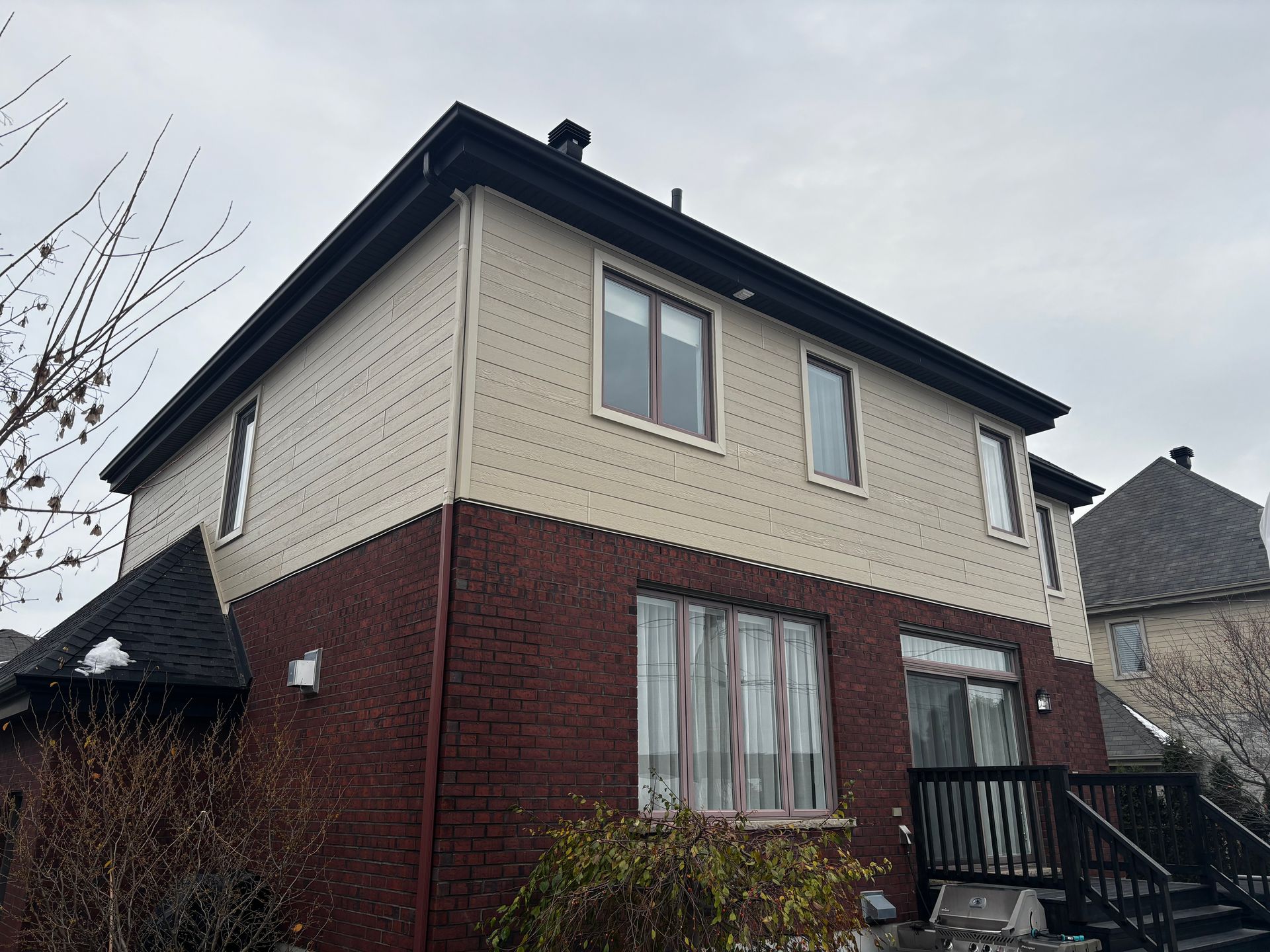 Maison à deux étages avec soubassement en briques, bardage beige et toit noir, sous un ciel nuageux.