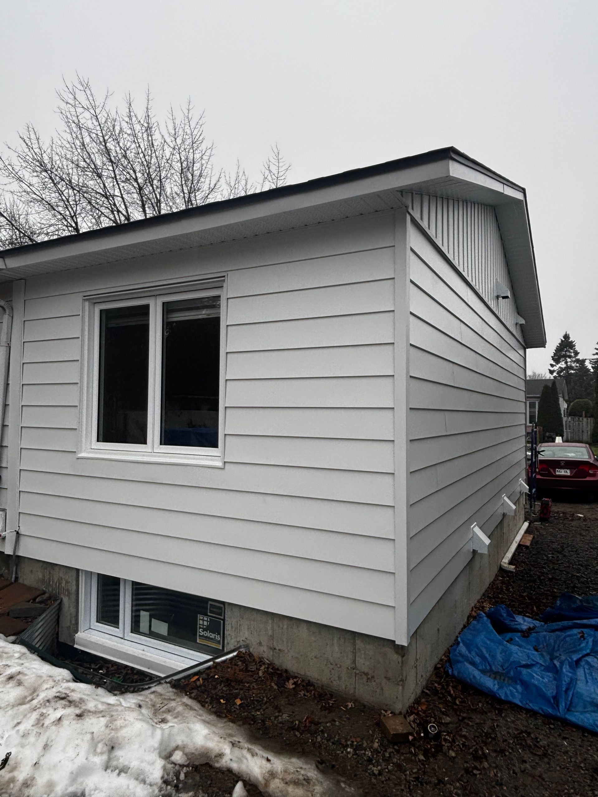 Extérieur d'une extension de maison avec bardage et fenêtres blancs, sur des fondations en béton, sous la neige et un ciel couvert.