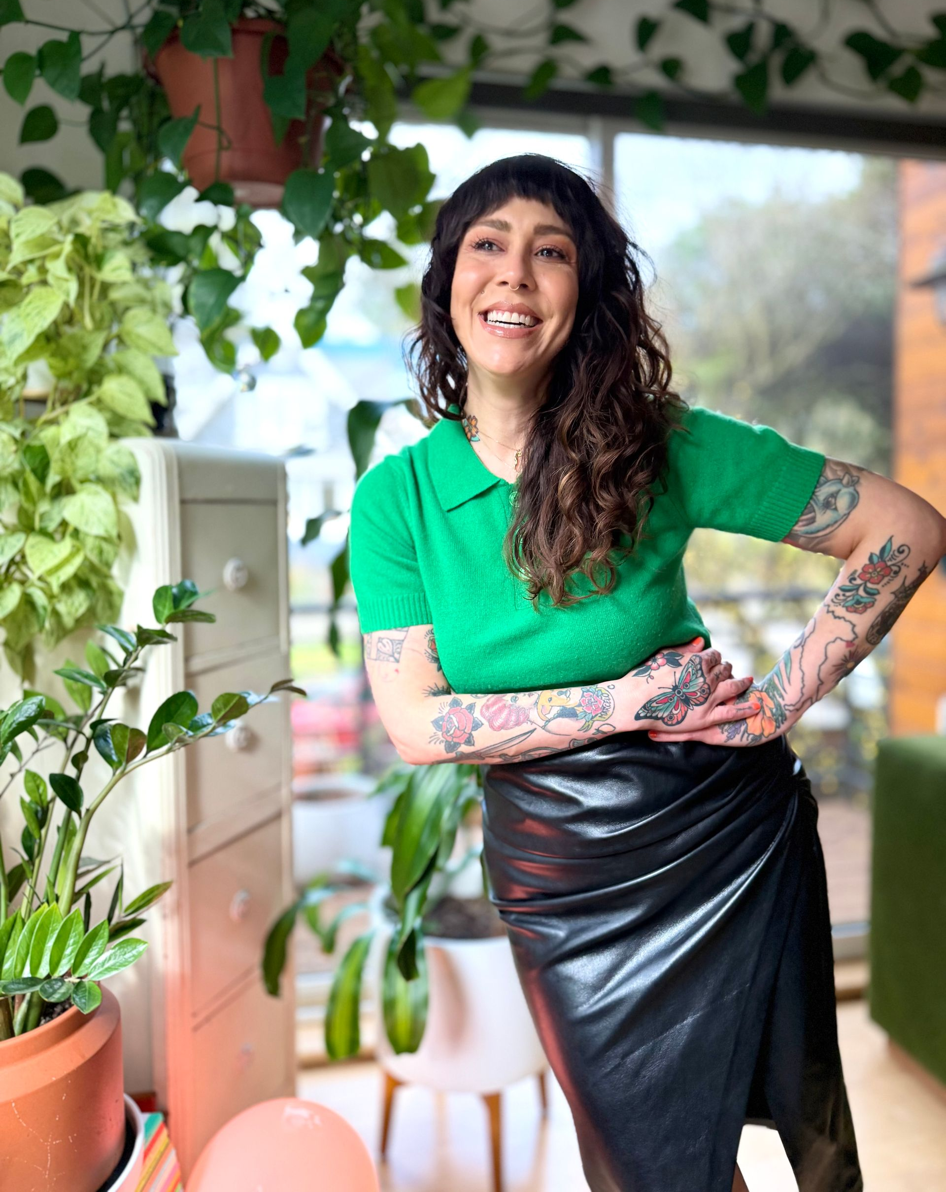 Woman smiling, posing with hands on hips, in a green top and black skirt, surrounded by plants.