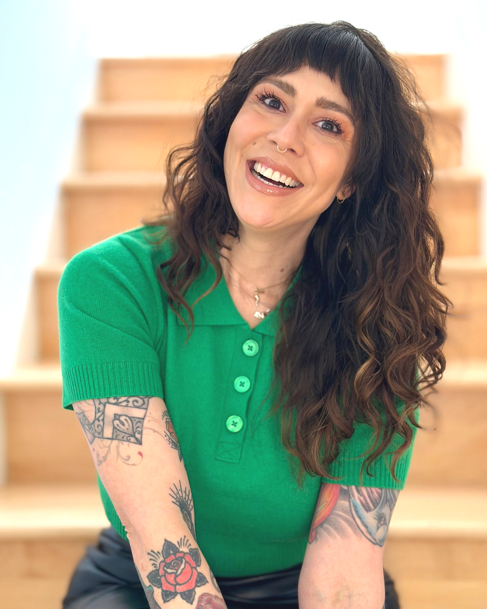 Woman with dark curly hair and bangs, wearing a gray sweater, smiling, with a nose ring and dark lipstick.