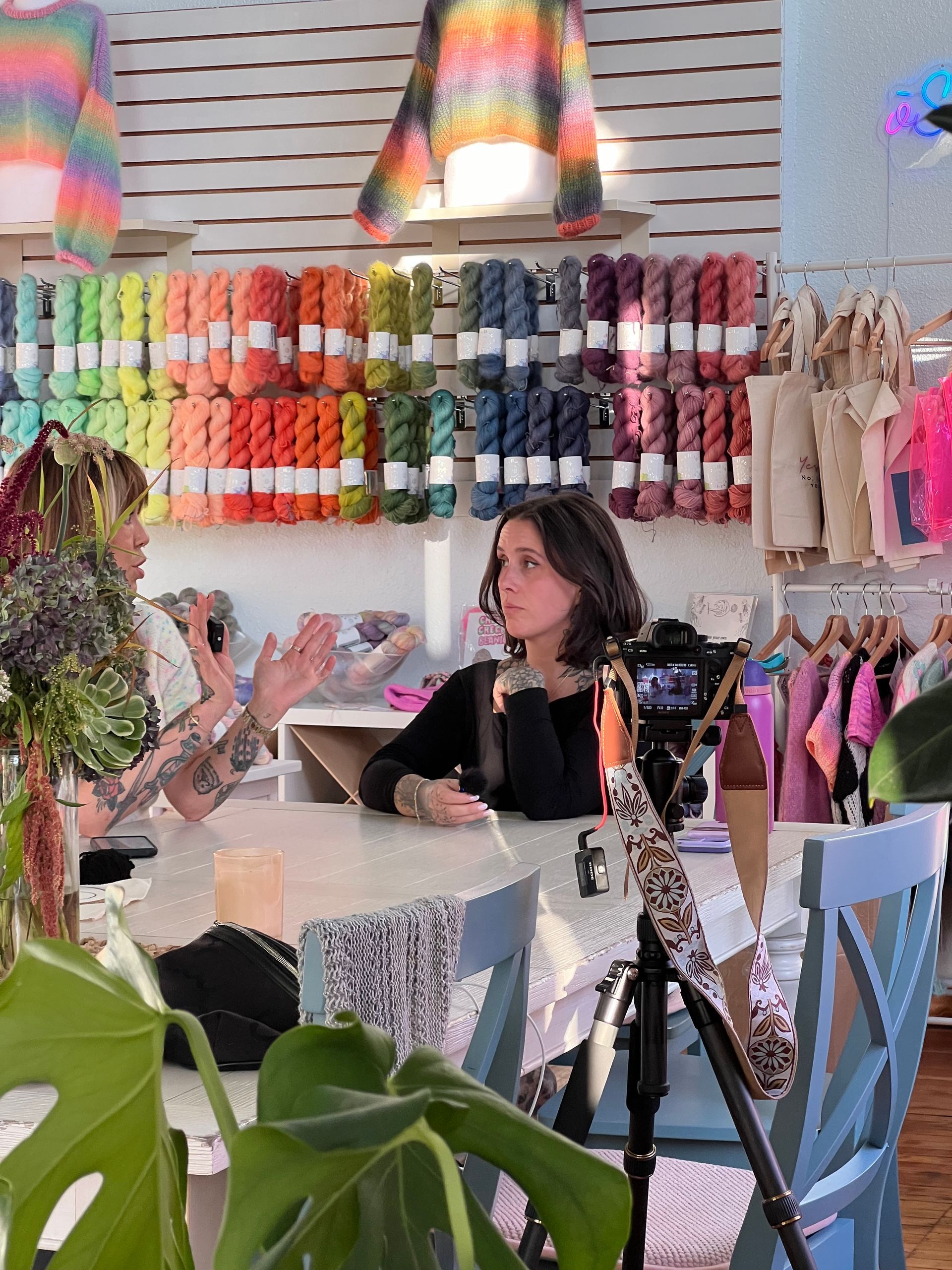 Two women in a yarn shop filming with a camera. Colorful yarn and sweater are on display.