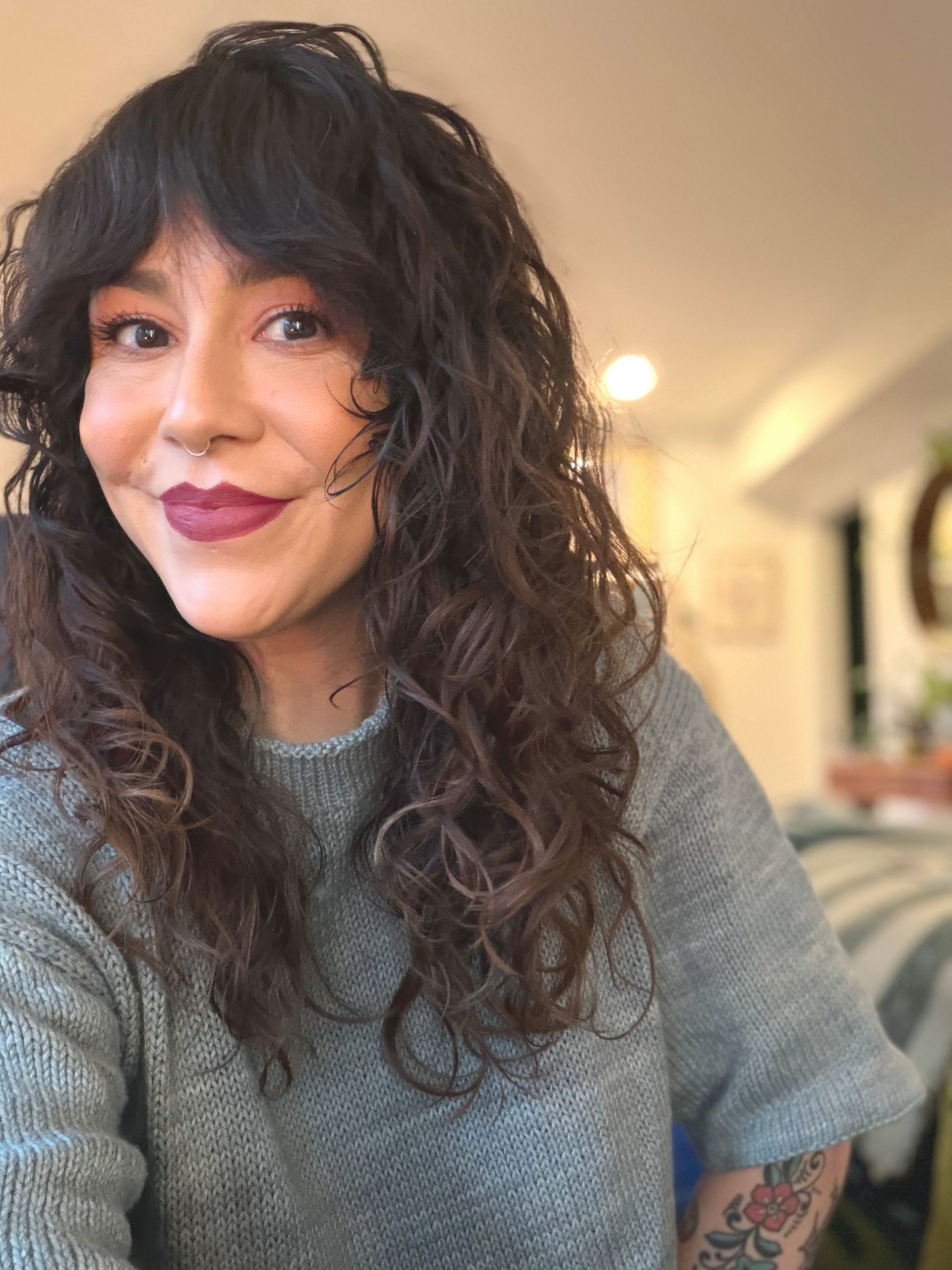 Woman with dark curly hair and bangs, wearing a gray sweater, smiling, with a nose ring and dark lipstick.