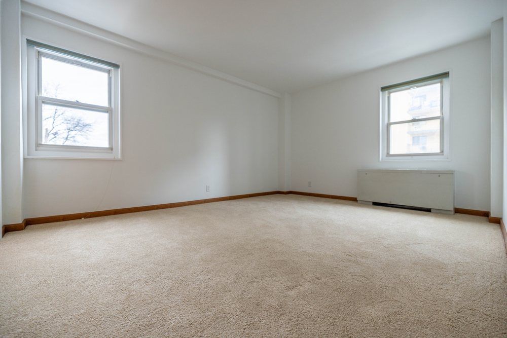 Empty beige-carpeted room with white walls and two windows.