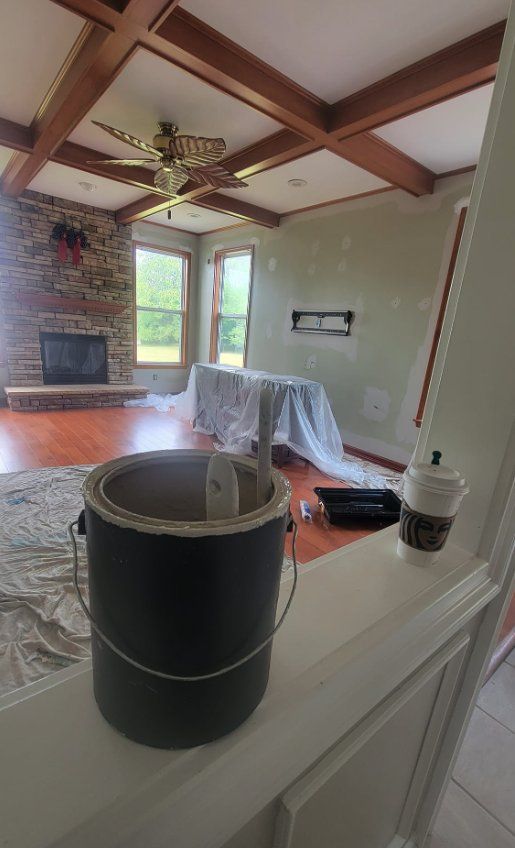 Living room with exposed wood beams, ceiling fan, fireplace, and paint supplies on a covered counter.