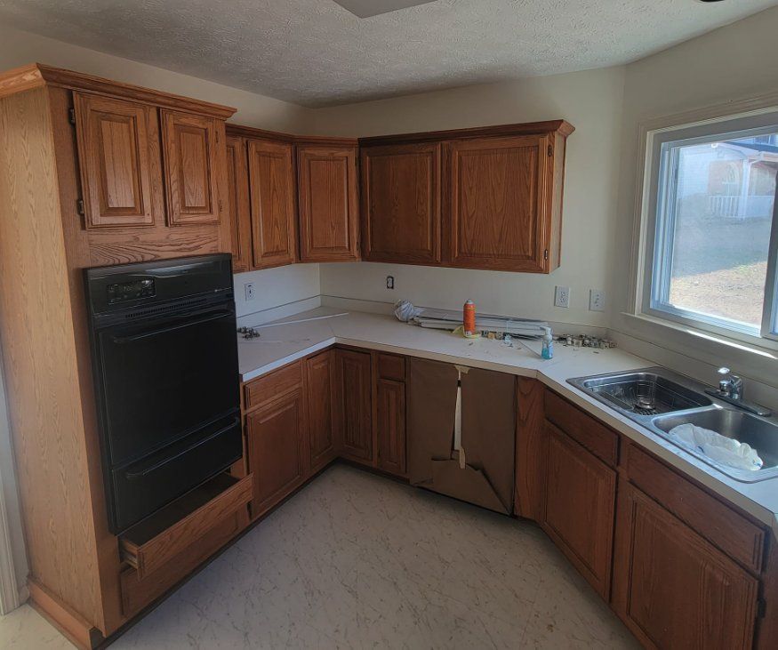 Compact L-shaped kitchen with wood cabinets, black oven, white counters, and a sink by a bright window