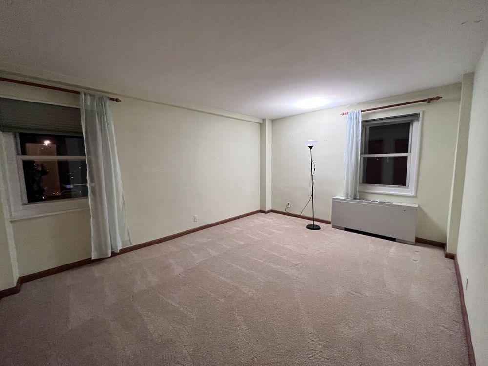 Empty beige living room with carpet, two windows, and a floor lamp near the far wall