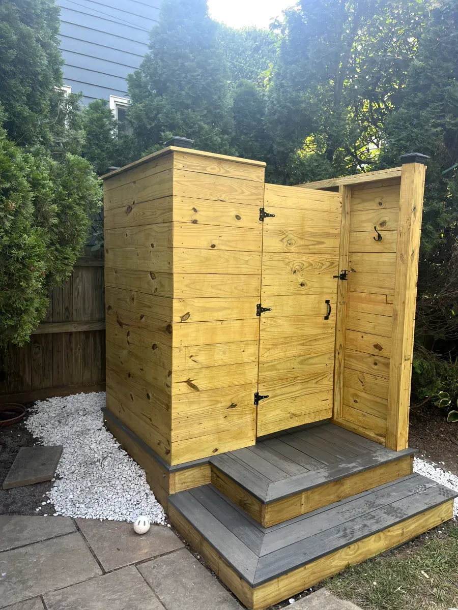 Wooden outdoor shower with gray steps surrounded by white pebbles, in a backyard setting.