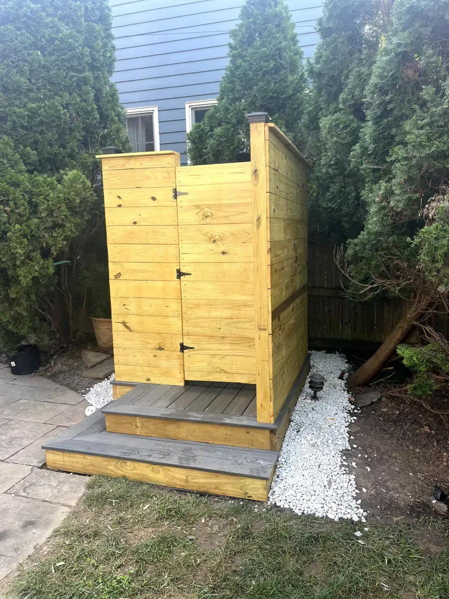 Wooden outdoor shower with steps, painted yellow, set in a backyard with landscaping.