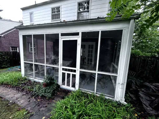 Screened porch attached to a two-story house with a white door, windows, and green foliage.
