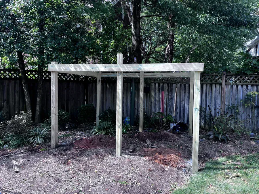 Wooden pergola in a backyard, built with posts, beams, and a top structure. Surrounded by dirt, trees, and a fence.