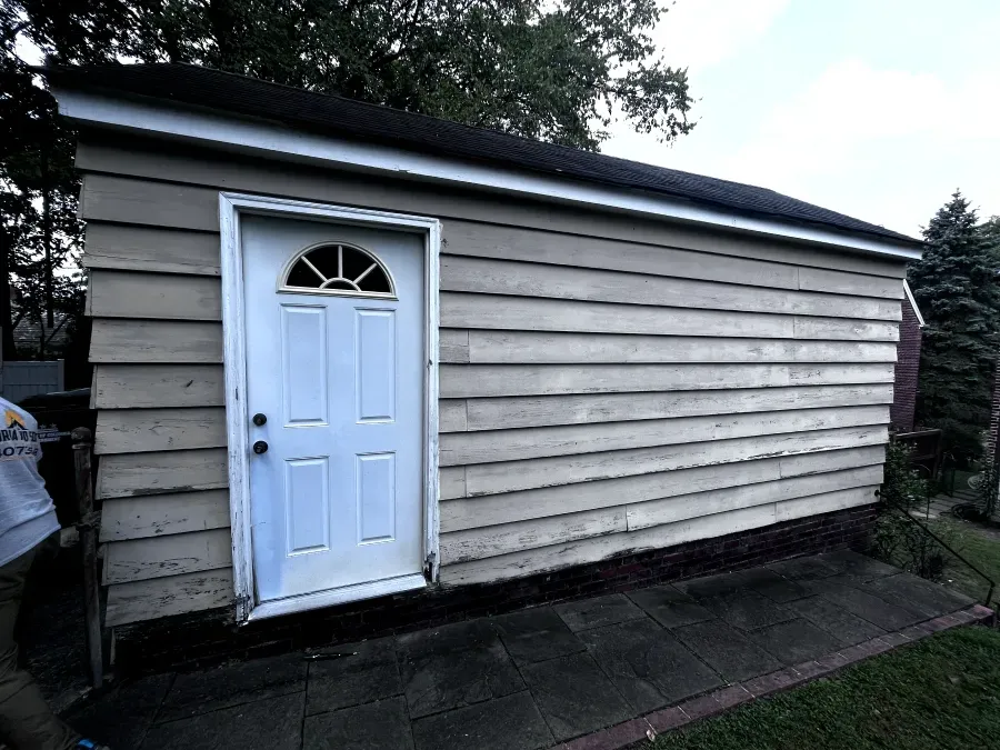 Small, beige, rectangular building with white door and black trim. Set on a dark paved area with greenery.