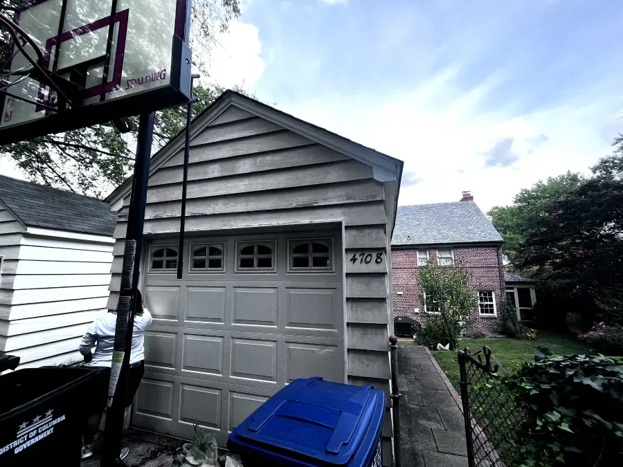 Garage with a basketball hoop, blue trash bin, and a house in the background under a cloudy sky.