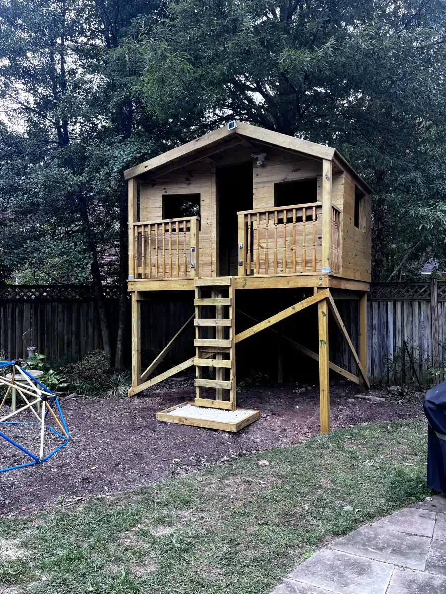 Wooden treehouse on stilts in backyard with ladder leading up.