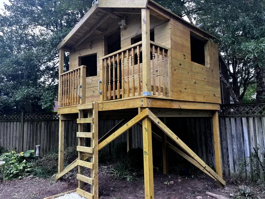 Wooden treehouse with a ladder, in a backyard with a wooden fence.
