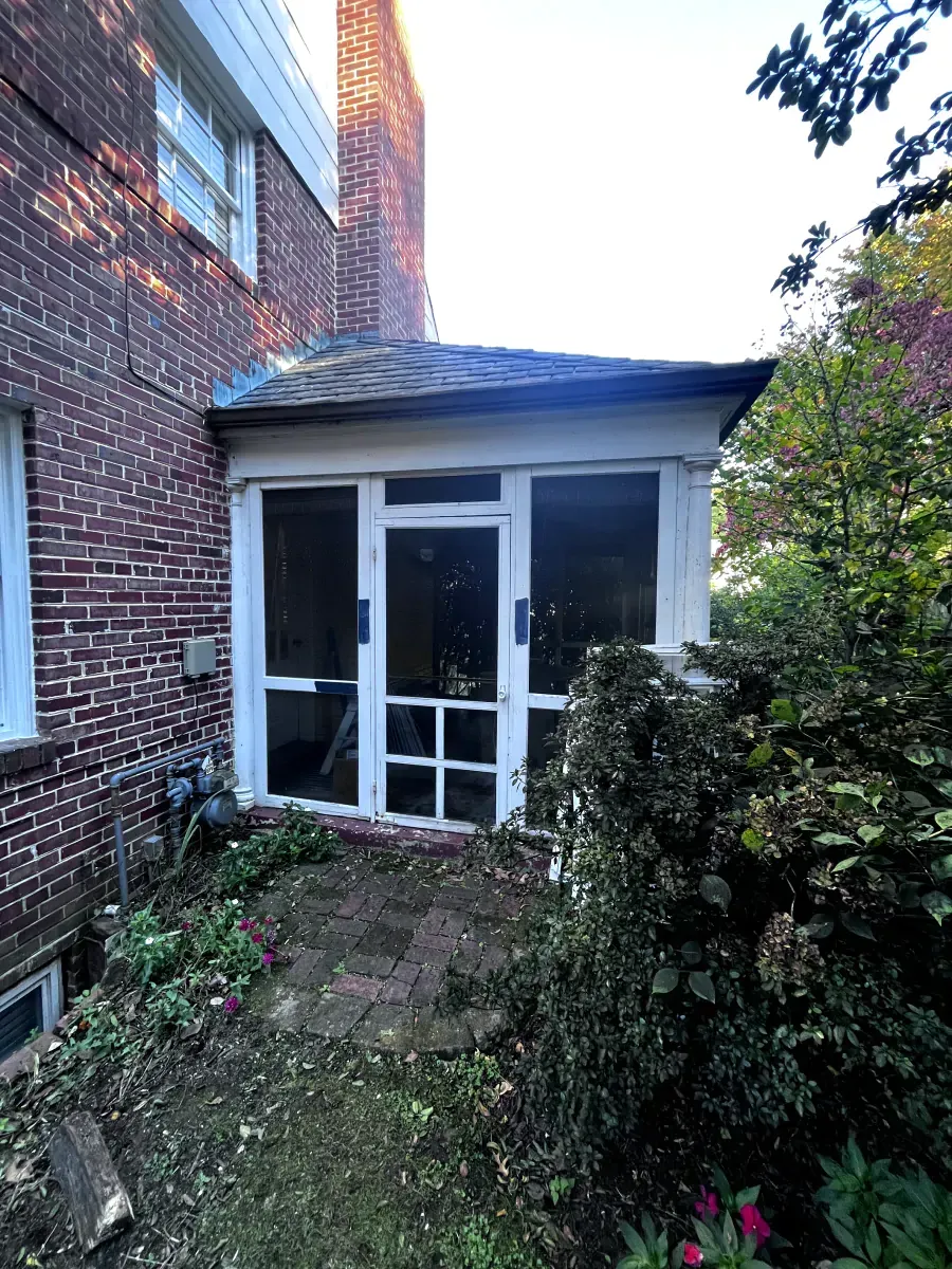 Screened porch attached to a brick house, surrounded by plants and a garden.