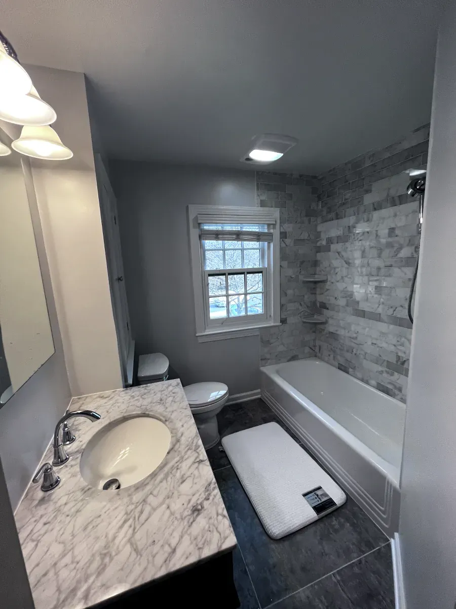 Bathroom with gray walls, marble vanity, white tub, and stone tile shower surround.