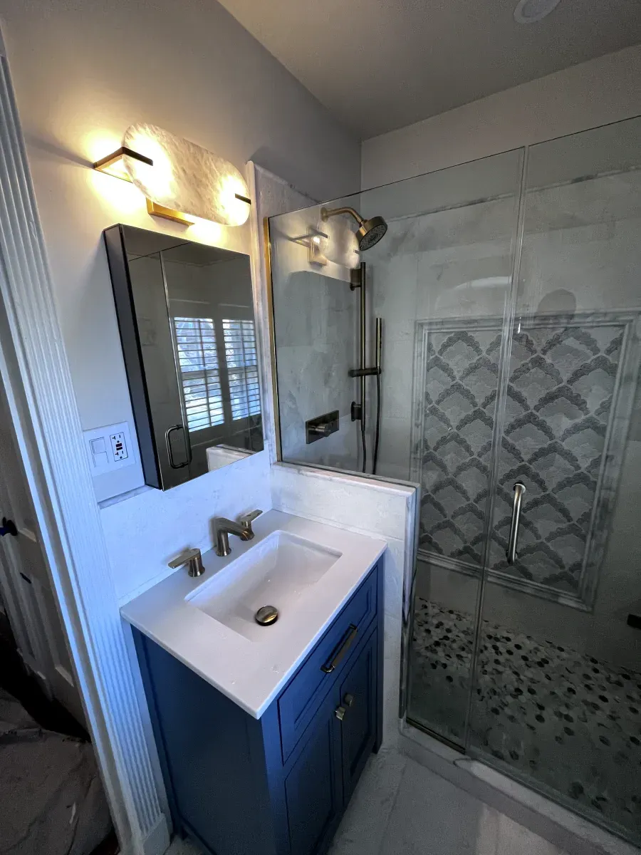 Bathroom with blue vanity, white sink, glass shower, gray and white tiles, and gold fixtures.