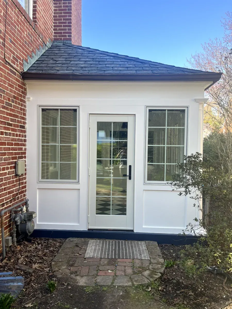 White-framed enclosed porch with glass door and windows, on a brick house. Brown roof. Brick pathway.