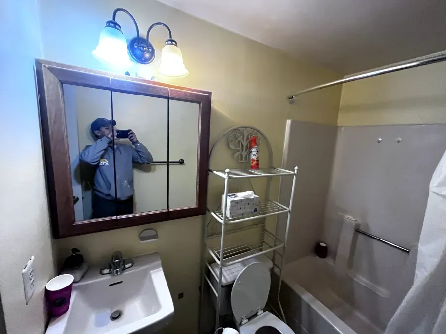 Person taking photo in a small bathroom. Mirror, sink, toilet, shelves, and tub are visible.