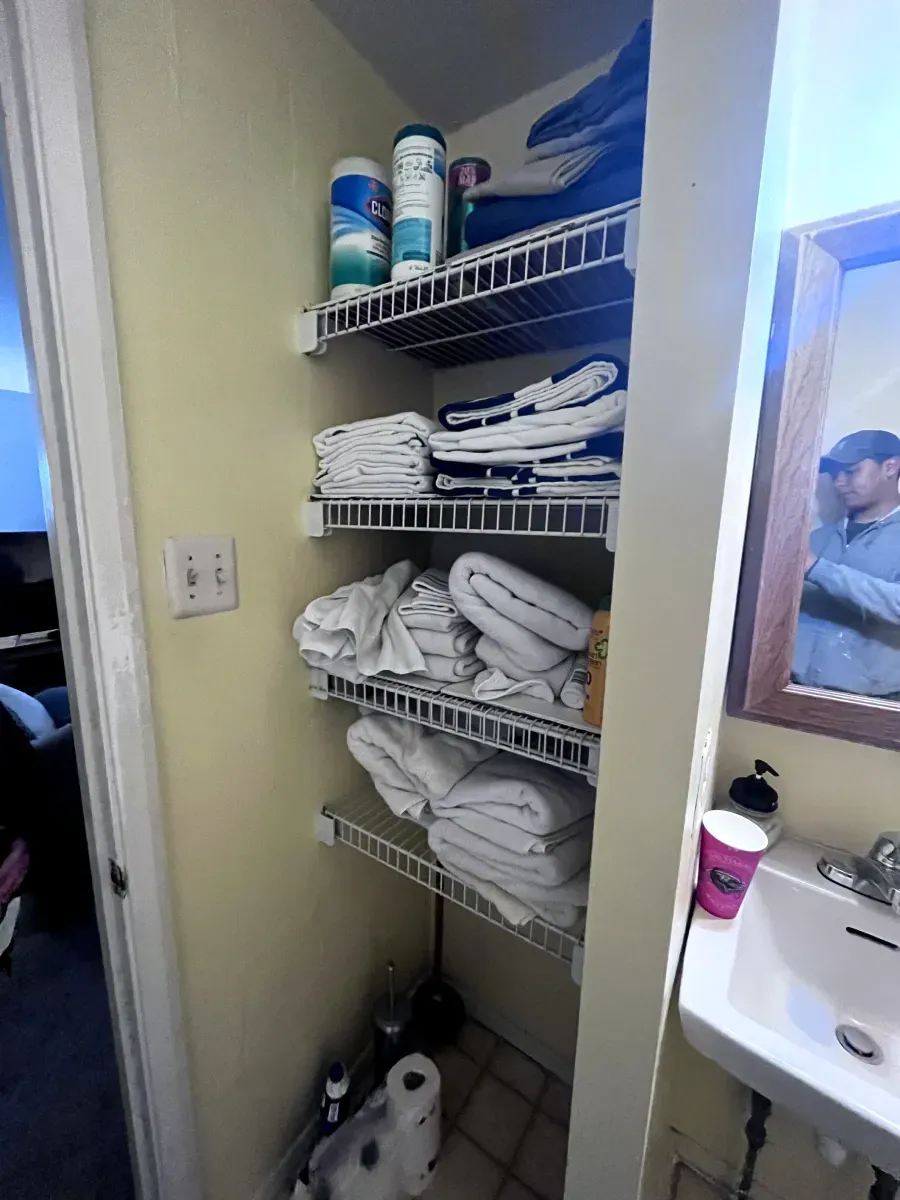 Small bathroom with wire shelving holding folded towels and cleaning supplies.