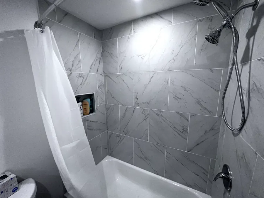Gray-tiled shower with a white curtain and a built-in shelf. Silver showerhead and faucet are visible.