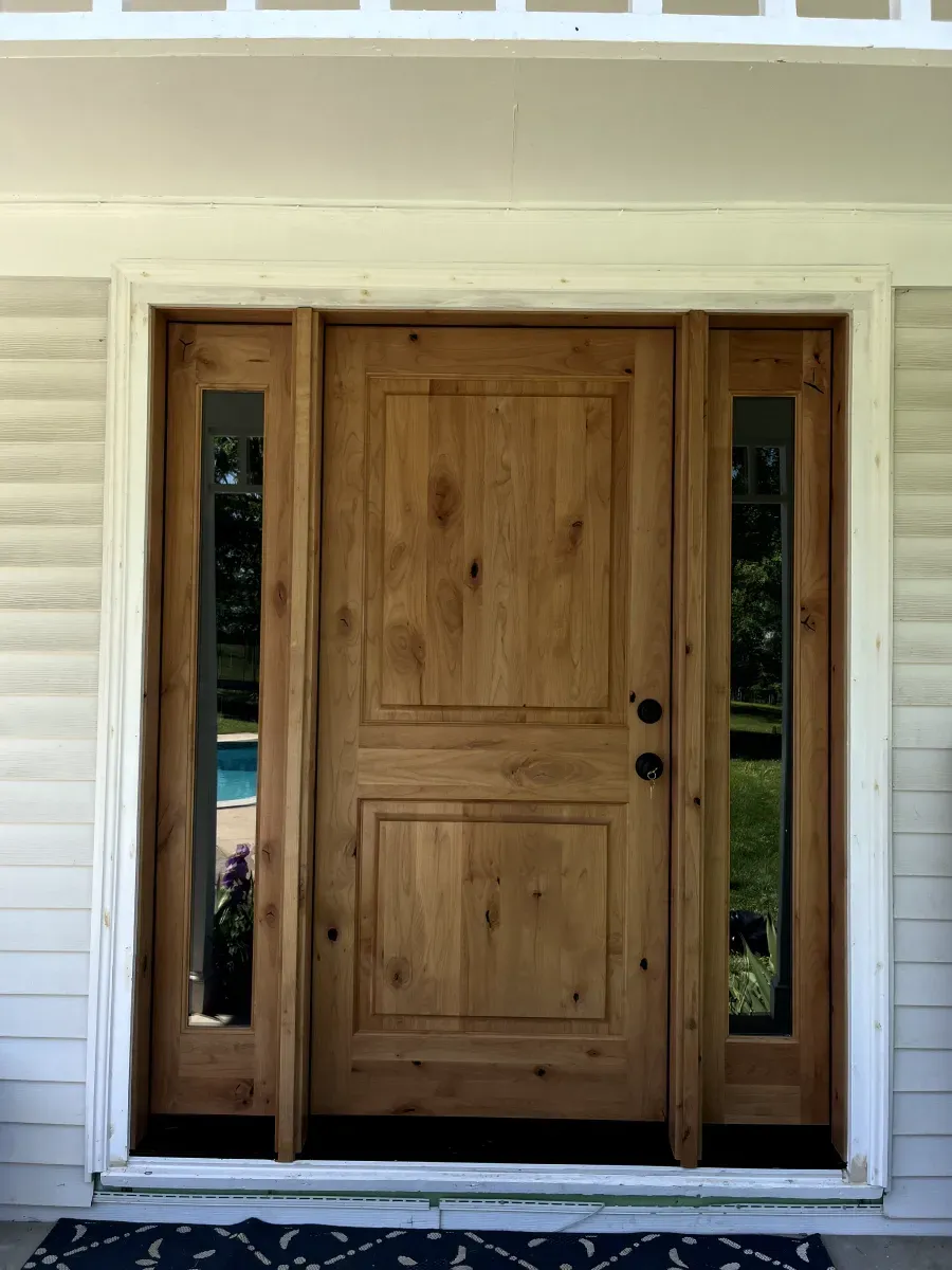 Wooden front door with side glass panels; light wood color, white trim, dark doormat.