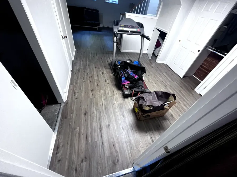 Narrow hallway with treadmill, bags, and cardboard box on wood-look flooring.