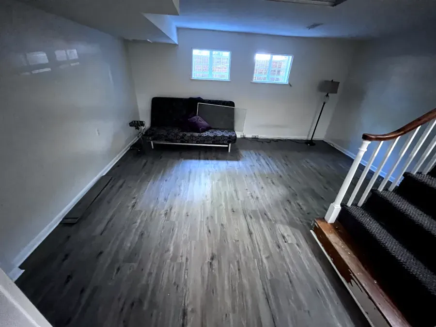 Basement room with gray wood-look flooring, a futon, small windows, and stairs leading up.