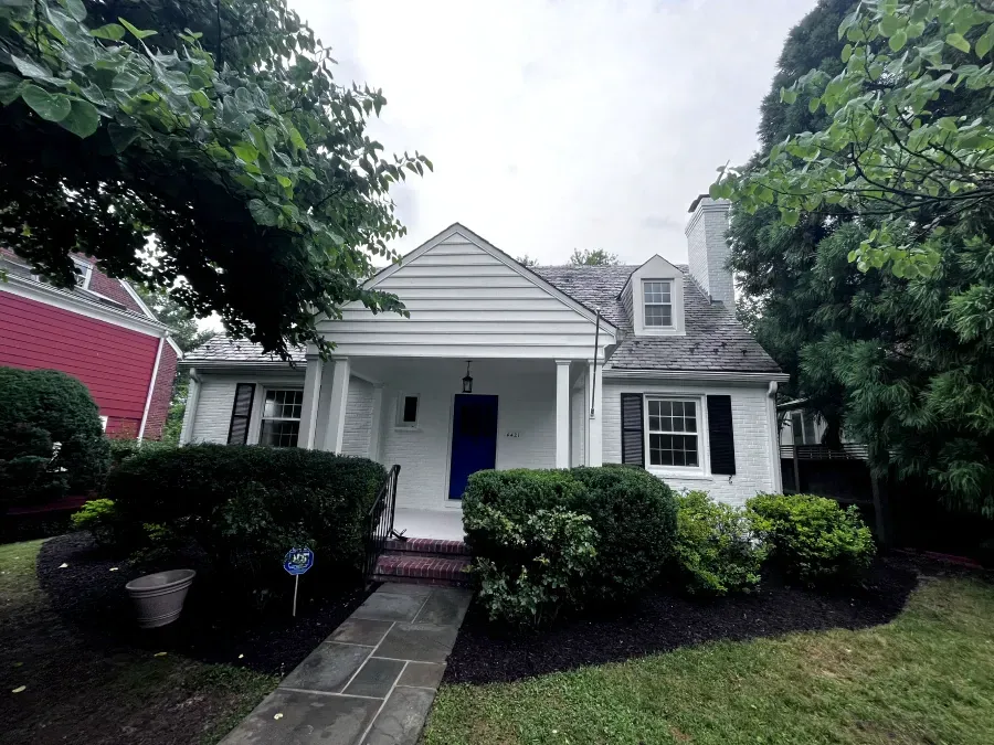 White cottage house with blue door, black shutters, and lush greenery in front.