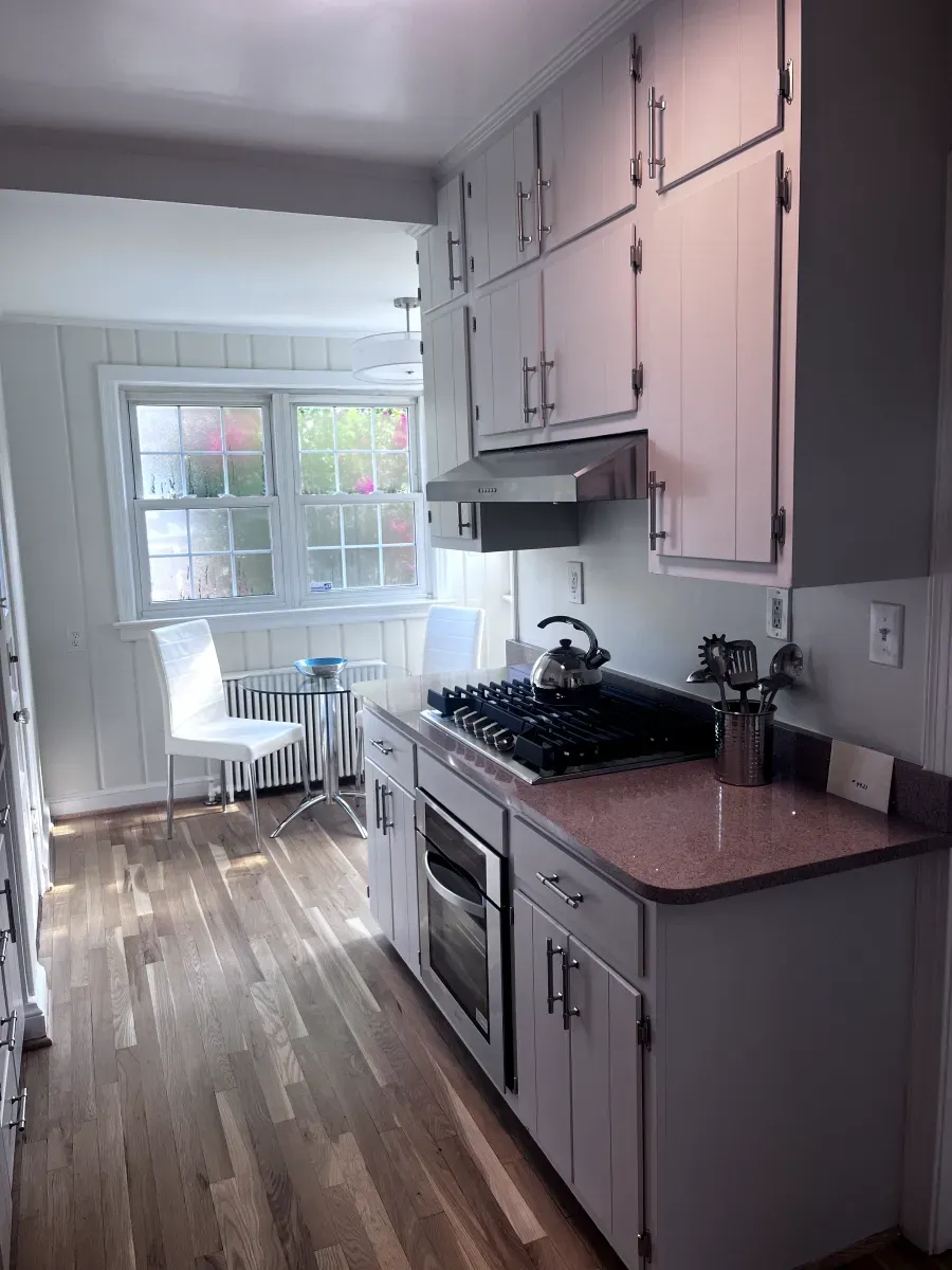 A narrow kitchen with white cabinets, gas range, and a window with a table and chair.