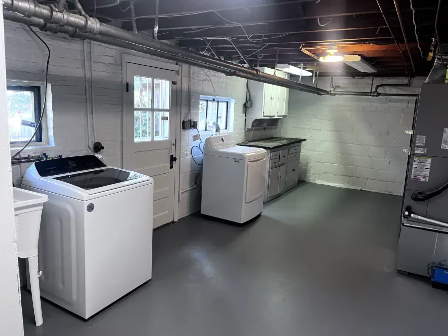 Laundry room with white appliances, gray floor, brick walls, and pipes.